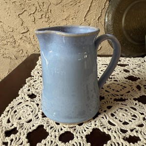 May include: A light blue ceramic pitcher with a handle. The pitcher has a slightly textured surface and a light brown rim. It is sitting on a white, lacy doily.