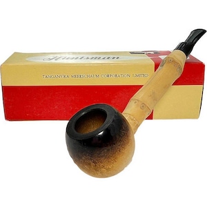 May include: A Huntsman pipe with a bamboo stem and a dark brown bowl. The pipe rests in front of a box with the name "Huntsman" and "TANGANYIKA MEERSCHAUM CORPORATION LIMITED" printed on it. The box has a red and yellow color scheme.