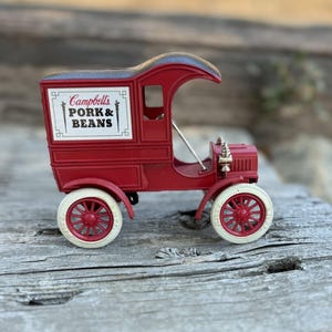 May include: A red vintage toy truck with white tires and red spokes. The truck has a boxy back with the words "Campbell's PORK & BEANS" on the side. The truck is a miniature replica of an older model.
