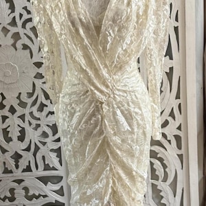 May include: Ivory lace dress with long sleeves and a V-neck. The dress features a ruched bodice and a fitted silhouette, displayed on a wooden dress form. The background includes a white, carved wooden screen.