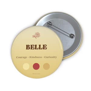 May include: A round, cream-colored button with the name "BELLE" in brown text. Below the name are the words "Courage, Kindness, Curiosity." A small rose graphic is above the name. The button has a pin back.