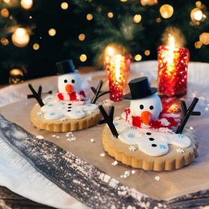 Puede incluir: Dos galletas con forma de muñeco de nieve con sombreros de copa negros, narices de zanahoria y bufandas a rayas rojas y blancas. Las galletas están sobre una superficie blanca con un fondo borroso de luces y velas navideñas.