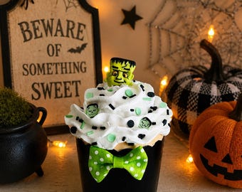 May include: A Halloween-themed cupcake with white frosting, green and black sprinkles, and a Frankenstein head topper. The cupcake sits in a black cup with a green polka-dot bow tie. A sign reads "Beware of Something Sweet."