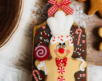 May include: A gingerbread man cookie decorated with white icing, a chef's hat, and a red bow tie. The cookie is on a wooden cutting board with a red and white checkered bow. Candy canes and sprinkles add to the festive design.
