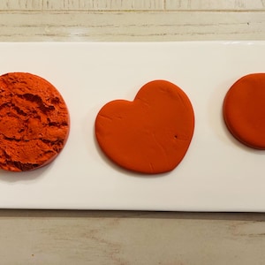 Puede incluir: Una bandeja rectangular blanca muestra tres formas de arcilla naranja: un círculo texturizado, un corazón y un círculo liso. Las formas están dispuestas en una fila sobre un fondo claro de vetas de madera.