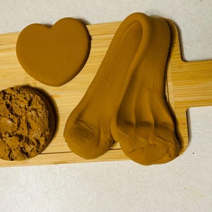 May include: A wooden serving board with a heart-shaped object, a round object, and two long, curved objects. All objects are brown. The board has a handle with a hole.