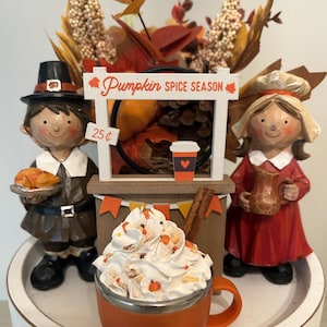 May include: A decorative fall-themed display featuring a pumpkin spice latte with whipped cream, a sign that reads "Pumpkin Spice Season", and figurines of a pilgrim and a girl. The latte is in an orange mug with a cinnamon stick.