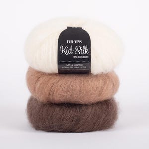 Peut inclure: Trois pelotes de laine empilées les unes sur les autres. La pelote du haut est blanche, la pelote du milieu est brun clair et la pelote du bas est brun foncé. L'étiquette sur la pelote du haut indique "DROPS Kid-Silk UNI COLOUR Soft & luxurious in Super Kid Mohair & Silk."