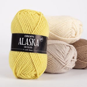 May include: Four skeins of Drops Alaska yarn in different colors: yellow, cream, beige, and brown. The yarn is 100% wool and is labeled "Drops Alaska Uni Colour - A wool classic".
