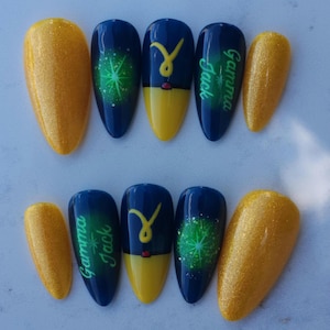May include: A set of almond-shaped false nails. The nails feature a glittery gold finish and a design with navy blue, yellow, and green accents. Some nails have the text "Gamma Jack".