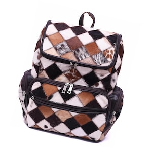 Peut inclure: Un sac à dos patchwork avec un motif géométrique de carrés marron, noirs et blancs. Le sac est doté d'une poignée supérieure, d'une poche avant zippée et de deux poches latérales. Le sac à dos est fait d'une matière texturée.