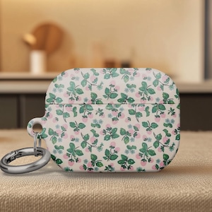 May include: A white AirPod case with a floral pattern of green leaves and pink flowers. The case has a silver carabiner clip attached. The background is blurred, suggesting a tabletop setting.