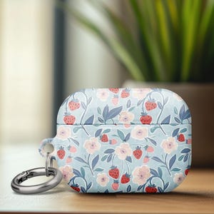 May include: A light blue AirPod case with a strawberry and floral pattern. The case features a repeating design of red strawberries, pink flowers, and green leaves. A silver-colored carabiner clip is attached to the case.