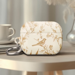 May include: Cream-colored AirPod case with a gold bird and floral design. The case has a silver carabiner clip attached. A white teacup and saucer are in the background, along with a vase of dried flowers.