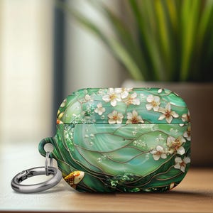May include: Green and white floral AirPod case with a silver carabiner clip. The case features a design of white flowers, gold accents, and a koi fish. The case is on a wooden surface.