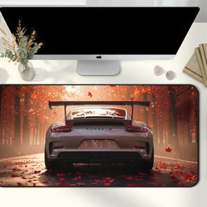May include: A large rectangular desk mat featuring a silver sports car on a road lined with red autumn trees. The word "PORSCHE" is visible on the car. The mat is surrounded by office supplies, including a computer monitor, notebooks, and a phone.