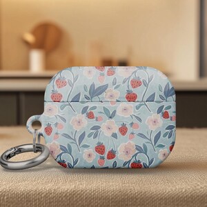 May include: A light blue AirPod case with a floral and strawberry pattern. The case has a silver-colored carabiner clip attached. The design features pink flowers, red strawberries, and green leaves.