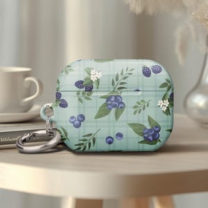 May include: A light blue AirPod case with a pattern of blueberries, blackberries, and white flowers. The case has a silver carabiner clip attached. The design is on a plaid background.