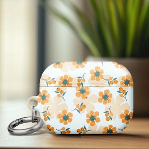 May include: White AirPods case with a floral pattern of orange and yellow flowers with teal centers. The case has a silver carabiner clip attached. The background includes a wooden surface and a potted plant.
