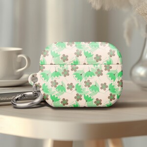 May include: A floral patterned AirPod case in shades of green and brown on a cream background. The case has a silver carabiner clip attached. A white coffee cup and a book are in the background.