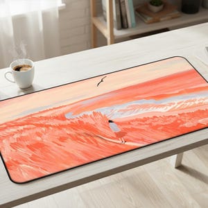 May include: Rectangular desk mat featuring a watercolor painting of a person walking on a beach at sunset. The artwork is predominantly orange and pink, with a black border. A seagull flies in the sky. A cup of coffee and a succulent plant are on the table.