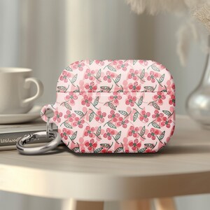 May include: A pink AirPod case with a floral pattern featuring red flowers and green leaves. The case has a silver carabiner clip attached. A white coffee cup and a book are in the background.