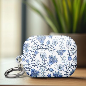 May include: White and blue floral patterned AirPods Pro case with a silver carabiner clip. The case features a detailed design of blue flowers and vines on a white background. The case is on a wooden surface.
