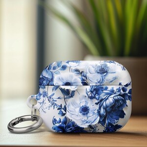 May include: White and blue floral patterned AirPod case with a silver carabiner clip. The case features a detailed design of blue flowers and foliage on a white background. The case is sitting on a wooden surface.