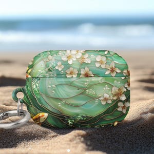May include: Green and white floral AirPods Pro case with a gold fish design. The case has a silver carabiner clip and is set on a sandy beach with the ocean in the background. The case features white flowers and bubbles.
