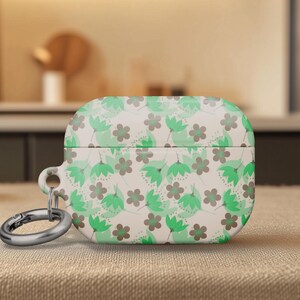 May include: A floral patterned AirPod case in shades of green and gray on a cream background. The case has a silver carabiner clip attached. The design features stylized flowers and leaves. The case is sitting on a textured surface.