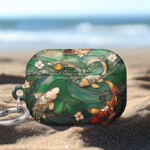 May include: A decorative case for wireless earbuds, featuring a stained-glass design of koi fish, lily pads, and flowers in shades of green, gold, and orange. The case has a silver carabiner clip attached. The background is a sandy beach with the ocean in the distance.