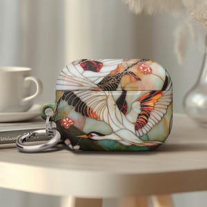 May include: AirPods Pro case with a crane design. The case features a colorful illustration of white cranes with orange and black accents, set against a backdrop of green and peach tones. A silver carabiner clip is attached.
