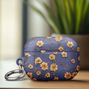 May include: A blue case for wireless earbuds, adorned with a pattern of gold-colored flowers. The case has a silver carabiner clip attached. The design gives a floral and elegant look to the tech accessory.