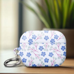 May include: White AirPod case with a floral pattern of blue flowers and pink stems. The case has a silver carabiner clip attached. The background includes a wooden surface and a blurred view of a plant.