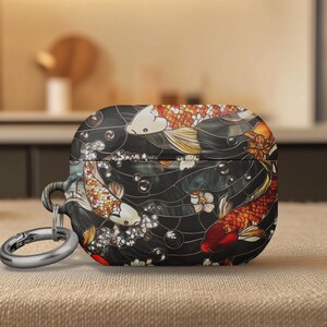 May include: A black AirPod case featuring a koi fish design in shades of orange, red, and white. The case has a silver carabiner clip. The design includes water, bubbles, and floral accents.