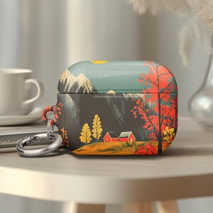 May include: A decorative case for wireless earbuds featuring a landscape design. The case has a dark gray base with a colorful illustration of a mountain range, trees, and red houses. A silver carabiner clip is attached.