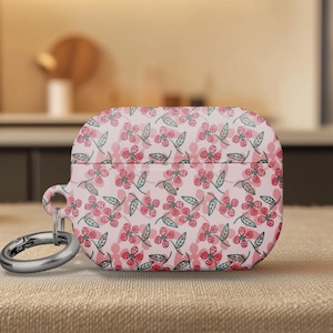 May include: A light pink AirPod case with a repeating floral pattern in red and green. The case has a silver metal ring attached for easy carrying. The background is blurred, showing a kitchen setting.