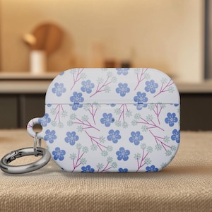 May include: White AirPod case with a floral pattern of blue flowers and pink stems. The case has a silver carabiner clip attached. The background is blurred, suggesting a home or office setting.