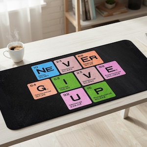 Könnte beinhalten: Schwarze Schreibtischunterlage mit dem Schriftzug "NEVER GIVE UP" in Elementen des Periodensystems. Die Unterlage hat einen schwarzen Hintergrund mit bunten Quadraten. Eine Kaffeetasse und eine kleine Pflanze sind auf dem Tisch.