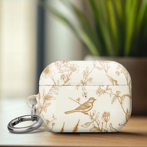 May include: White AirPod case with a gold floral and bird design. The case has a silver carabiner clip attached. The design features a bird perched on a branch, surrounded by flowers and foliage. The case is sitting on a wooden surface.