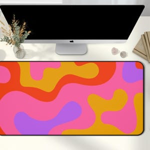 May include: A rectangular desk mat with a vibrant, retro-inspired design. The mat features a black border and a colorful pattern of swirling shapes in shades of pink, orange, yellow, and purple. The mat is placed on a white desk.