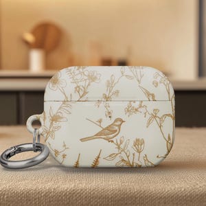 May include: A white AirPod case with a bird and floral design. The case features a detailed illustration of a bird perched on a branch, surrounded by delicate botanical elements in a warm, earthy tone. A silver carabiner is attached.
