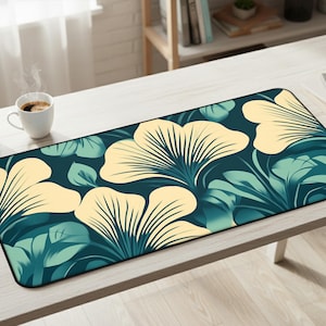 May include: A rectangular desk mat with a botanical design. It features large, cream-coloured fan-shaped flowers and teal leaves against a dark teal background. The mat is on a light-coloured wooden desk.