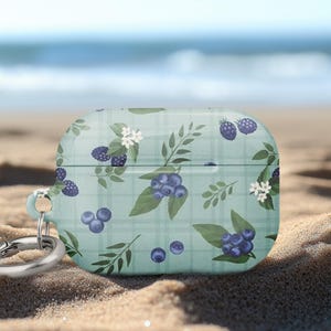 May include: A light blue case for wireless earbuds, adorned with a pattern of blueberries, blackberries, white flowers, and green leaves. The case has a silver carabiner clip. The background is a sandy beach with the ocean in the distance.