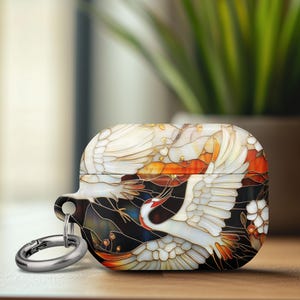 May include: AirPods case featuring a crane design. The case has a white, orange, and black stained glass effect. A silver carabiner clip is attached. The design includes white cranes, orange accents, and white flowers.