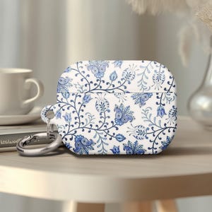 May include: White and blue floral patterned AirPod case with a silver carabiner clip. The case is rectangular and sits on a light-colored wooden surface. A white teacup and saucer are in the background.