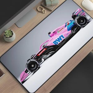 May include: A rectangular desk mat featuring a high-resolution graphic of a Formula 1 race car. The car is primarily pink, black, and blue, with visible branding including BWT, Alpine, Castrol, and Mapfre. The design is set against a white background.