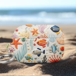 May include: A white case for wireless earbuds, adorned with colorful embroidered sea creatures. The design includes orange starfish, blue and orange fish, and various sea plants. A silver carabiner clip is attached. The case is resting on sand with the ocean in the background.