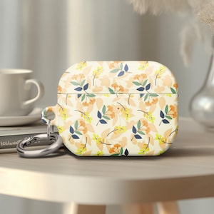 May include: A floral patterned AirPods case with a silver carabiner clip. The case features a cream background with a repeating pattern of orange, yellow, and blue flowers and leaves. The case is sitting on a light-colored wooden surface.