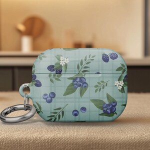 May include: A teal AirPod case with a pattern of blueberries, leaves, and white flowers. The case has a silver-colored carabiner clip attached. The design is on a plaid background.